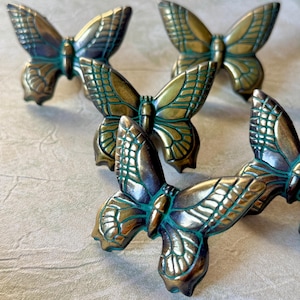 May include: Close-up of several bronze butterfly-shaped cabinet pulls. The butterfly wings have a detailed, textured design with a teal-colored patina. The pulls are arranged on a neutral-colored surface.