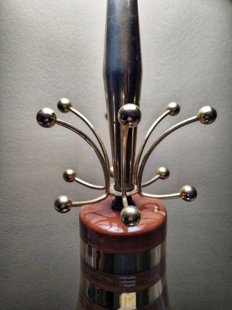 K&ouml;nnte beinhalten: Eine Vintage-Lampe mit braun-goldenem Sockel, silbernem Schaft und goldenen Kugelakzenten. Das Design der Lampe erinnert an den Mid-Century-Modern-Stil. Der Sockel hat einen Durchmesser von ca. 15 cm.