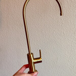May include: A gold-colored kitchen faucet with a curved spout and a single lever handle. The faucet is made of metal and has a modern design. The faucet is held by a hand.