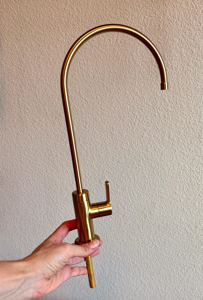 May include: A gold-colored kitchen faucet with a curved spout and a single lever handle. The faucet is made of metal and has a modern design. The faucet is held by a hand.