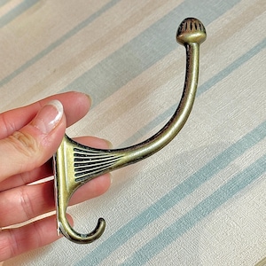 May include: Antique brass-colored wall hook with a curved design. The hook features a decorative, rounded top and a curved arm ending in a small hook. The base has a fan-like design with black lines. A vintage-style home decor item.