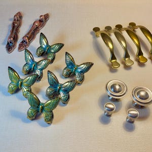 May include: A collection of decorative cabinet hardware, including bronze, brass, and silver-toned knobs and pulls. The selection features butterfly-shaped pulls with teal and gold accents, along with classic bar and knob designs.