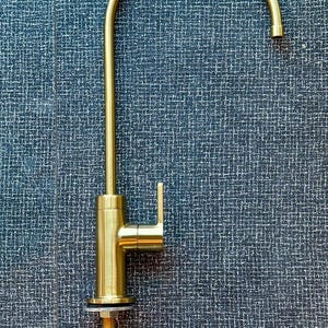 May include: A gold-colored kitchen faucet with a curved spout and a single lever handle. The faucet is mounted on a threaded rod and has a brushed finish. The background is a dark blue textured surface.