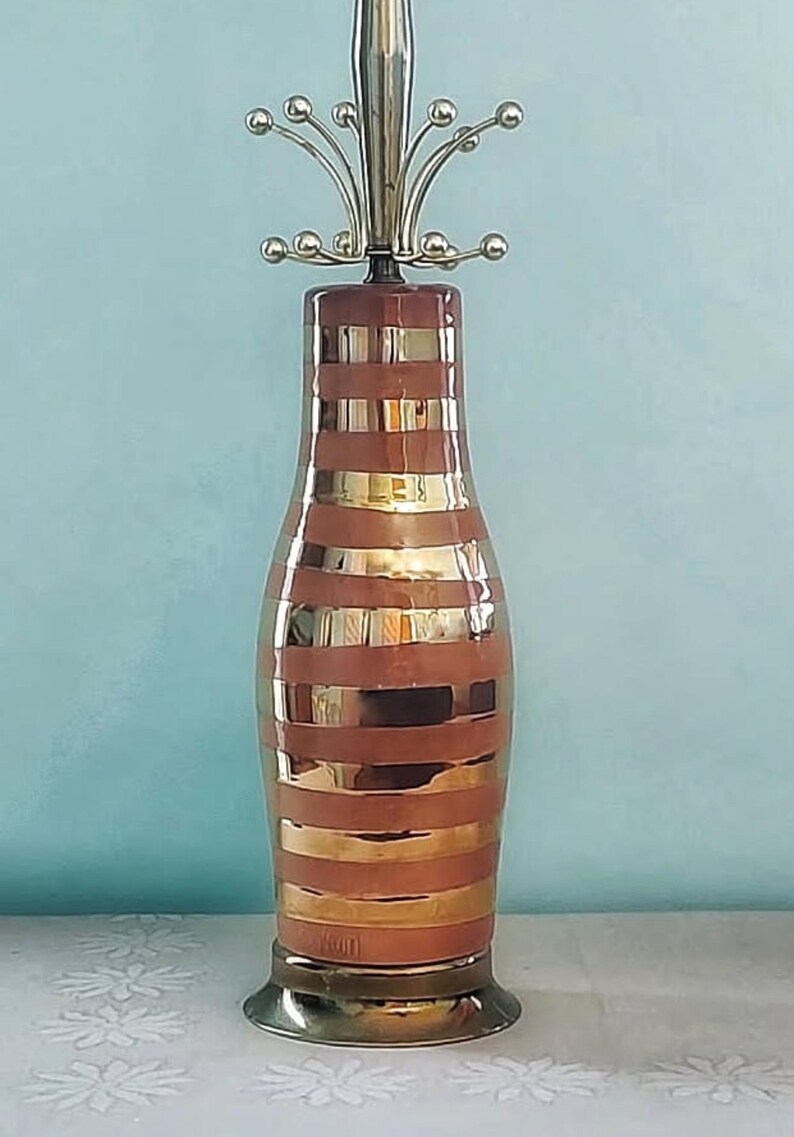 K&ouml;nnte beinhalten: Vintage Tischlampe mit einem braun-gold gestreiften Keramikk&ouml;rper. Die Lampe hat einen Messingfu&szlig; und eine dekorative Oberseite mit kleinen, runden Akzenten. Das Design der Lampe erinnert an die moderne Einrichtung der Mitte des Jahrhunderts.