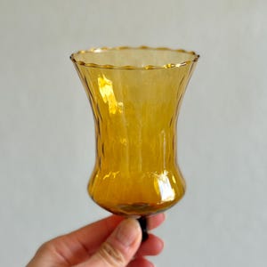May include: A hand-blown amber glass candle holder with a textured surface and a ruffled rim. The candle holder has a flared shape and a dark base. The glass is a warm, golden color.