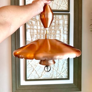 May include: A vintage copper pendant light fixture with a scalloped shade and a decorative ring. The fixture is held by a hand, showcasing its polished, reflective surface. The light has a diameter of approximately 18 inches.