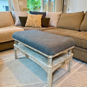 May include: A gray upholstered ottoman with a distressed white wooden base. The ottoman is rectangular and sits on a lower shelf. The room features a sectional sofa and patterned rug.