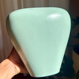 May include: A light green ceramic vase with a rounded top and a tapered base. The vase has a smooth, matte finish and is held by a person's hand. The background is blurred, with hints of a curtain and foliage.