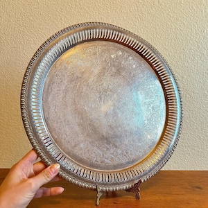 May include: Silver-toned, round serving tray with an ornate floral pattern. The tray has a decorative, ridged edge and is displayed on a small, decorative stand. The tray is held by a hand.