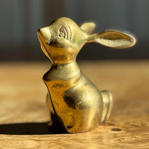 May include: A small, brass bunny figurine in a seated position. The bunny has long ears, a small tail, and detailed facial features. The brass has a polished, golden finish, and the figurine is set against a wooden surface.