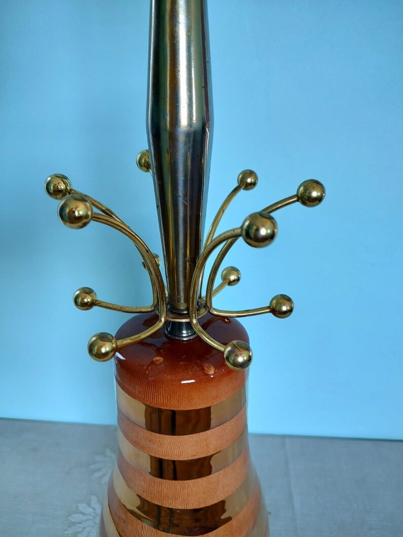 K&ouml;nnte beinhalten: Eine Tischlampe im Mid-Century-Modern-Stil mit einem Messingstiel und dekorativen Armen mit goldenen Kugeln. Der Lampensockel weist abwechselnd braune und goldene B&auml;nder auf. Ein Vintage-Dekorationsartikel.