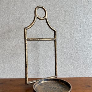 Könnte beinhalten: Ein Metallständer mit abgenutztem Gold-Finish, bestehend aus einer kreisförmigen oberen Schlaufe, einem rechteckigen Rahmen und einer runden Basisschale. Die Struktur besteht aus dünnen Metallstäben mit einer verwitterten Textur.