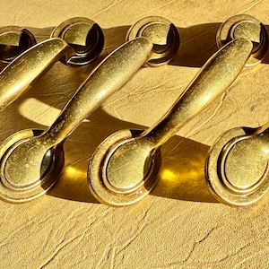 May include: A set of four gold-toned metal drawer pulls, each featuring a smooth, elongated handle attached to two circular mounting plates. The hardware has a polished finish with subtle signs of wear and is arranged in a diagonal row.