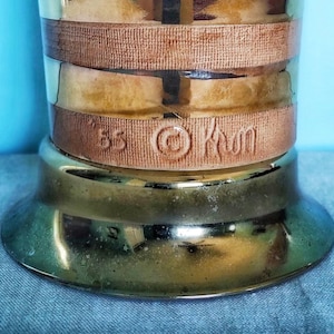 K&ouml;nnte beinhalten: Vintage Troph&auml;enfu&szlig; in Messingfarbe mit breitem, kreisf&ouml;rmigem Fu&szlig; und zylindrischem K&ouml;rper. Der K&ouml;rper hat zwei braune B&auml;nder und ein mittleres Band mit dem gepr&auml;gten Text "85 &copy; Kwm". Der Sockel hat eine gl&auml;nzende, reflektierende Oberfl&auml;che.