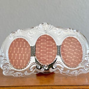 May include: A clear, ornate triple photo frame with an oval design. The frame has a decorative, swirling pattern around the edges. Each oval opening contains a patterned insert with a key motif. The frame sits on a wooden surface.