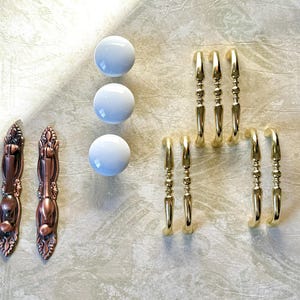 May include: An assortment of cabinet hardware, including three white ceramic knobs, several gold-toned handles, and two ornate bronze-colored pulls. The hardware is arranged on a textured, light-colored surface, showcasing various design styles.