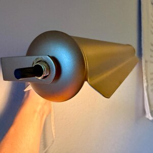 May include: A gold-colored wall-mounted lamp with a cylindrical body and a rectangular mounting bracket. The lamp has a directional design with a flared, wing-like shape. The lamp is turned on, emitting a warm, yellow light.
