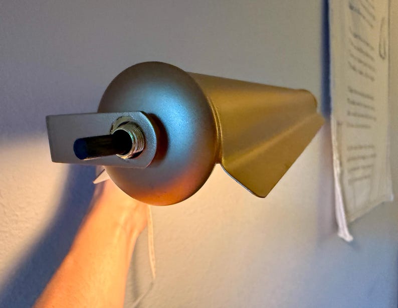 May include: A gold-colored wall-mounted lamp with a cylindrical body and a rectangular mounting bracket. The lamp has a directional design with a flared, wing-like shape. The lamp is turned on, emitting a warm, yellow light.