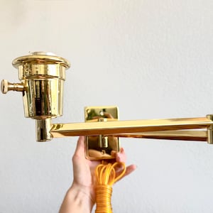 May include: A brass-toned wall-mounted lamp with an adjustable arm. The lamp features a cylindrical shade and a square mounting plate. A yellow tassel is visible.