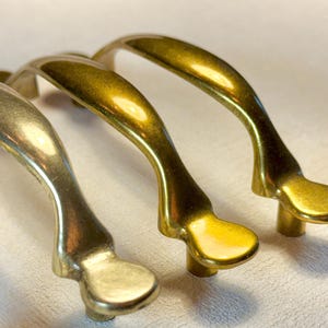 May include: Close-up of several brass cabinet handles. The handles have a curved design with a rounded base and a smooth, shiny finish. The brass color varies slightly between the handles, with some appearing more gold and others more silver.