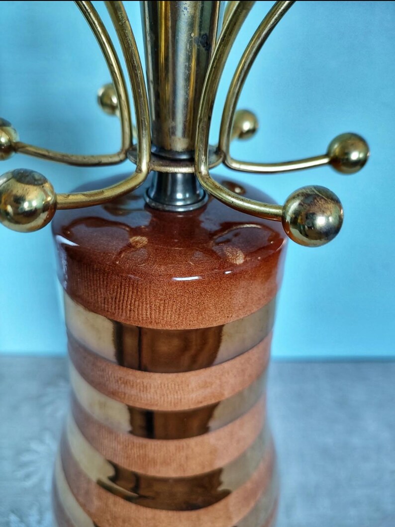 K&ouml;nnte beinhalten: Eine Vintage-Tischlampe mit einem braun-gold gestreiften Keramiksockel. Die Lampe hat einen Messingaufsatz mit gebogenen Armen und kugelf&ouml;rmigen Akzenten. Das Design der Lampe ist modern, mit einem gl&auml;nzenden Finish und einer Retro-&Auml;sthetik.