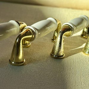 May include: Four cabinet pulls with a white ceramic center and gold-colored metal accents. The pulls have a curved design and are arranged in a row, casting shadows on the surface.