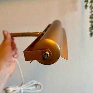 May include: A gold-colored, adjustable picture light with a cylindrical shade and a rectangular mounting bracket. The light is connected to a white electrical cord. The light is held by a hand.