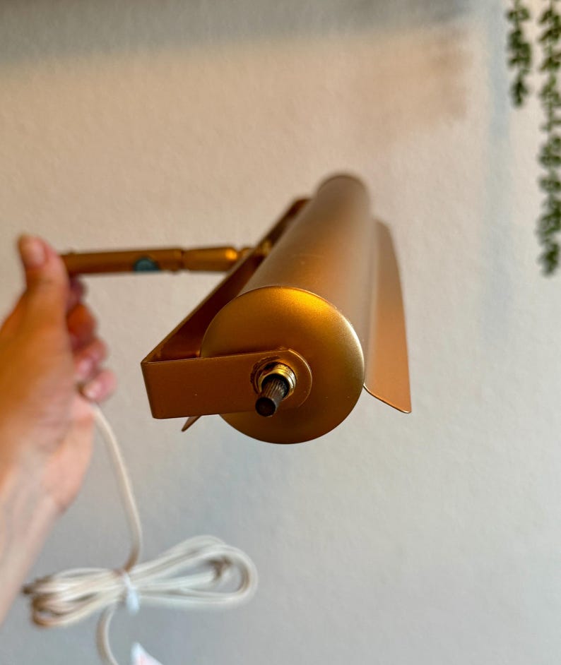 May include: A gold-colored, adjustable picture light with a cylindrical shade and a rectangular mounting bracket. The light is connected to a white electrical cord. The light is held by a hand.