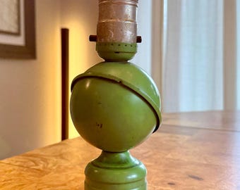 May include: A vintage green table lamp with a spherical shade and a round base. The lamp has a clear lightbulb that is lit. The lamp is made of metal and has a classic design. The lamp is sitting on a wooden table.