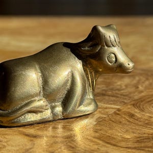 May include: A small, brass cow figurine in a resting position. The sculpture has a smooth, polished surface with visible details of the cow's features. The figurine is set against a warm, wooden background, with sunlight casting shadows.