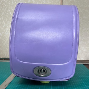 Puede incluir: Una mochila escolar tipo randoseru de color lavanda con un exterior liso y estructurado y una solapa superior redondeada. Un cierre giratorio de metal plateado está centrado en la parte inferior. Una regla de metal muestra un ancho de 28 cm.