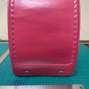 May include: A bright pink randoseru school backpack featuring a smooth, structured exterior with scalloped stitched edges and two small silver-toned metal bow accents at the bottom. A metal ruler shows the base width is approximately 29 cm.