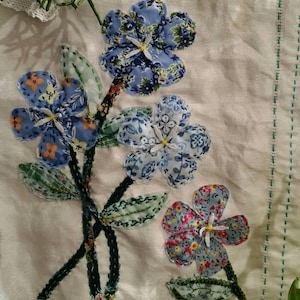 May include: A fabric floral arrangement featuring blue and red flowers with white centers and green leaves. The flowers are made from patterned fabric and are stitched onto a cream-colored background. The stems are dark green.