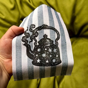 May include: A fabric patch with a black illustration of a teapot with stars on a blue and white striped background. The teapot has steam rising from the spout. The fabric is held against a green background.