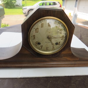 May include: A vintage wooden clock with a gold-coloured circular clock face. The clock has a dark brown wooden frame with an octagonal shape. The clock face displays black numerals and hands. The clock is sitting on a white surface.
