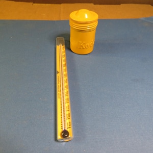 May include: A yellow Kodak film container and a yellow thermometer are displayed on a blue surface. The thermometer has black markings and a clear liquid inside. The film container has the Kodak logo.