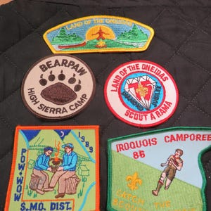 May include: Collection of embroidered patches with various designs and text. Includes a patch with a bear paw, one with a diamond, and others with camping and scouting themes. The patches are in various colors.