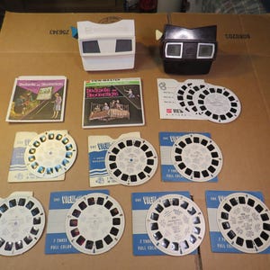 May include: A collection of vintage View-Master items, including two viewers in white and black, along with several reels and their packaging. The reels display colorful images, and the packaging features text and illustrations.