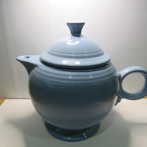 May include: A light blue ceramic teapot with a matching lid. The teapot has a rounded body, a curved spout, and a circular handle. The lid has concentric ridges and a small finial. The teapot sits on a pedestal base.