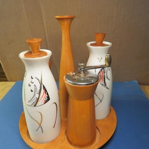 May include: A vintage kitchen set on a wooden tray. The set includes two white and gold bottles with fish designs, a wooden pepper grinder, and a tall wooden vase. The set has a mid-century modern aesthetic.