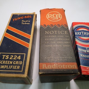 May include: Three vintage radio tube boxes. The boxes are labeled "Tung-Sol", "RCA Radiotron", and "Raytheon". The Tung-Sol box is navy blue with orange stripes and text. The RCA box is orange and black. The Raytheon box is red and blue.