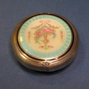 May include: A round, silver-toned compact with a floral design. The center features a bouquet of pink and blue flowers on a white background, surrounded by a light blue border. The compact has a black border and a detailed silver edge.