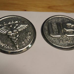 May include: Two circular silver-toned metal emblems. The left emblem features a caduceus symbol with the text U.S. ARMY MEDICAL CORPS. The right emblem features the letters U.S. with the word ARMY underneath, both set against a textured background.