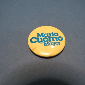 May include: A round, yellow button with the text "Mario Cuomo" in blue, above "For Mayor" in blue. The button is slightly worn and has a pin on the back. Political campaign memorabilia.
