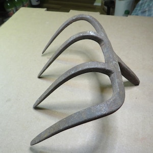 May include: A weathered, three-pronged metal tool with curved tines. The tool has a rustic, aged appearance, suggesting it is vintage or antique. The metal has a dark, mottled finish.