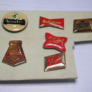 May include: A collection of six metal enamel pins featuring various beer brand logos including Heineken, Budweiser, Miller, Matt's, and Schlitz. The pins are arranged on a light-colored rectangular display card with gold-toned metal edges.