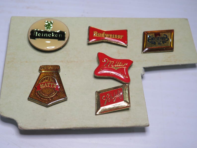 May include: A collection of six metal enamel pins featuring various beer brand logos including Heineken, Budweiser, Miller, Matt's, and Schlitz. The pins are arranged on a light-colored rectangular display card with gold-toned metal edges.