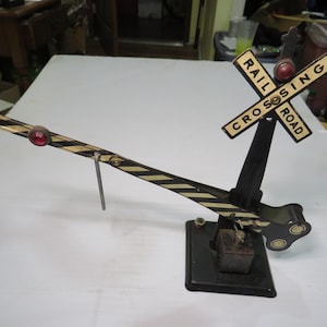 May include: Vintage model railroad crossing signal with a black base and arm. The arm has alternating black and yellow stripes and a red light. The sign reads "RAIL CROSSING ROAD".