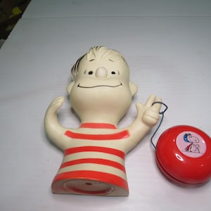 May include: A molded plastic figurine of a cartoon character with a round head and black hair, wearing a cream-colored shirt with horizontal red stripes. Beside the figurine is a round, glossy red yo-yo featuring a small illustration of a dog.