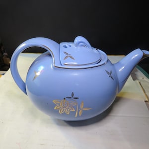 May include: A light blue ceramic teapot with a rounded body, a curved handle, and a spout. The lid features a small handle and decorative gold floral accents. Gold floral designs are also on the body.